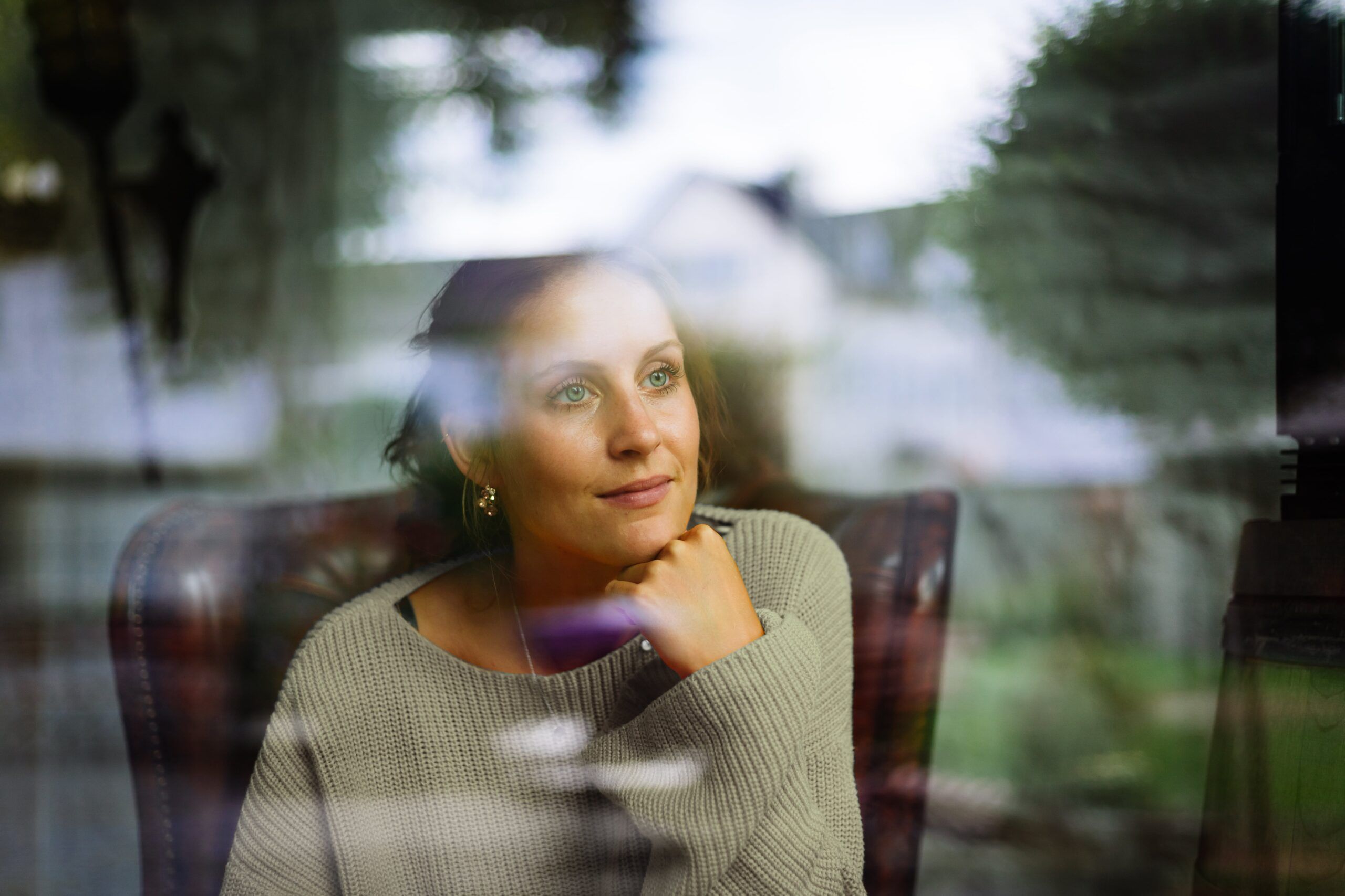 Een vrouw in een trui kijkt uit het raam, diep in gedachten verzonken, met de weerspiegelingen en het groen buiten.