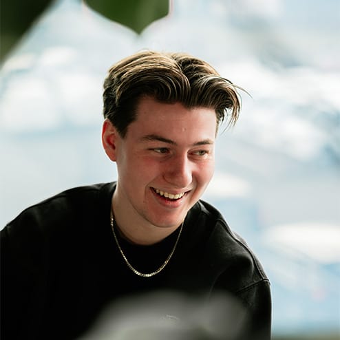 Lachende jongeman met bruin haar, zwart shirt en gouden ketting. De achtergrond is wazig.