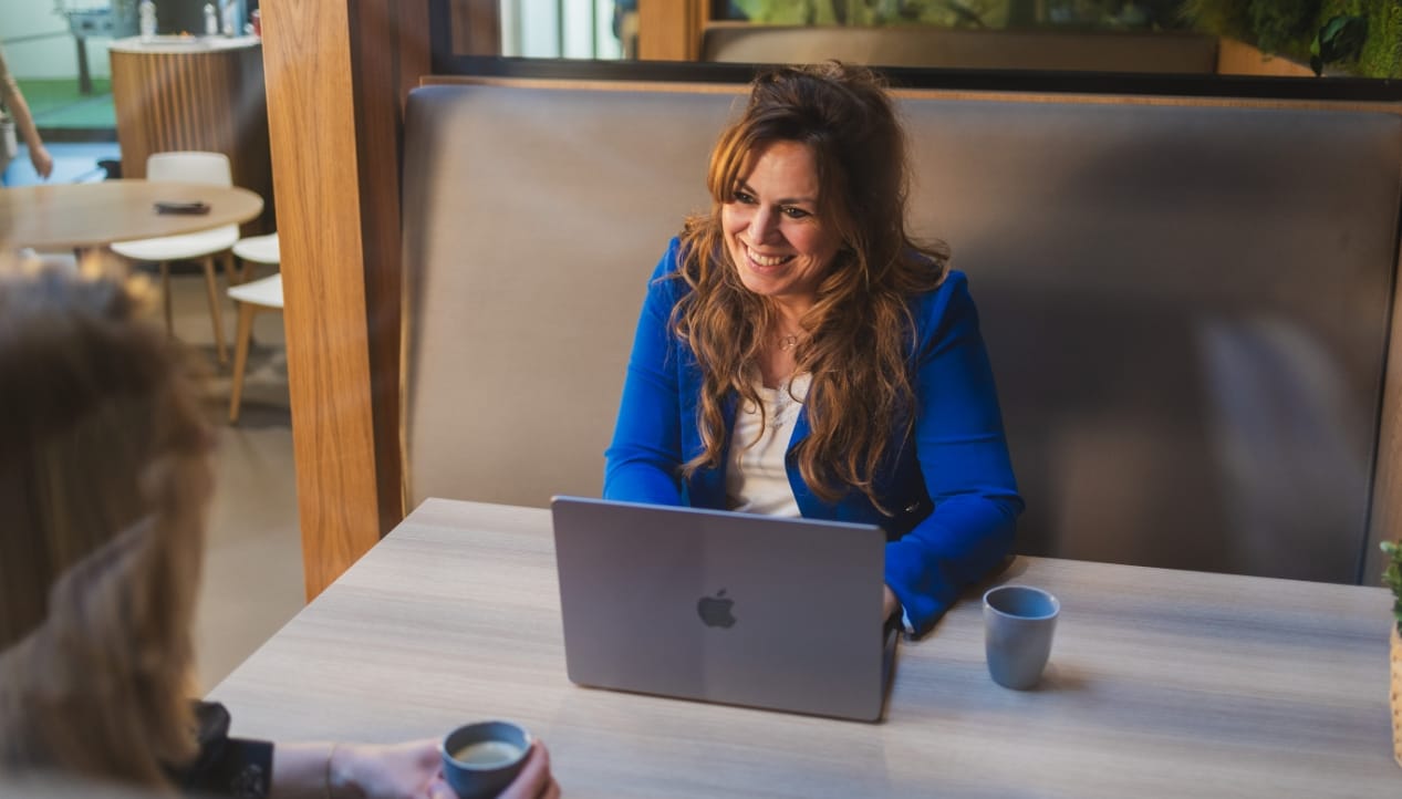 Een vrouw met een laptop en een HOBIJ-koffie glimlacht naar iemand in een café.