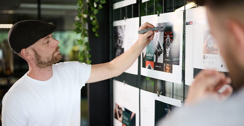 Een man met een pet is bezig met het aanpassen van de ontwerptekeningen voor een nieuw websiteproject dat aan de muur is geprikt.