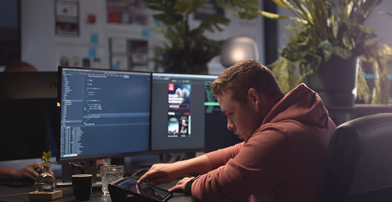 Persoon in hoodie verdiept in tablet, twee beeldschermen, weelderige planten; gefocust op 'website laten maken Roosendaal'.