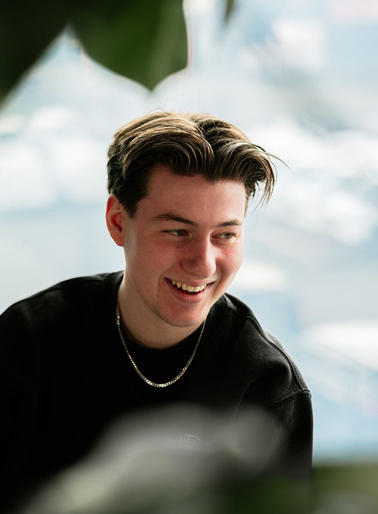 Jongeman met bruin haar lacht in een zwart shirt en een gouden ketting, tegen een onscherpe achtergrond.