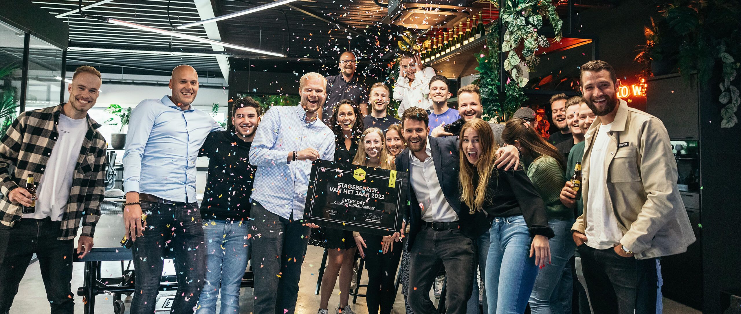 Mensen vieren het feest binnen met confetti en houden een ingelijst certificaat vast, te midden van planten en decoraties.