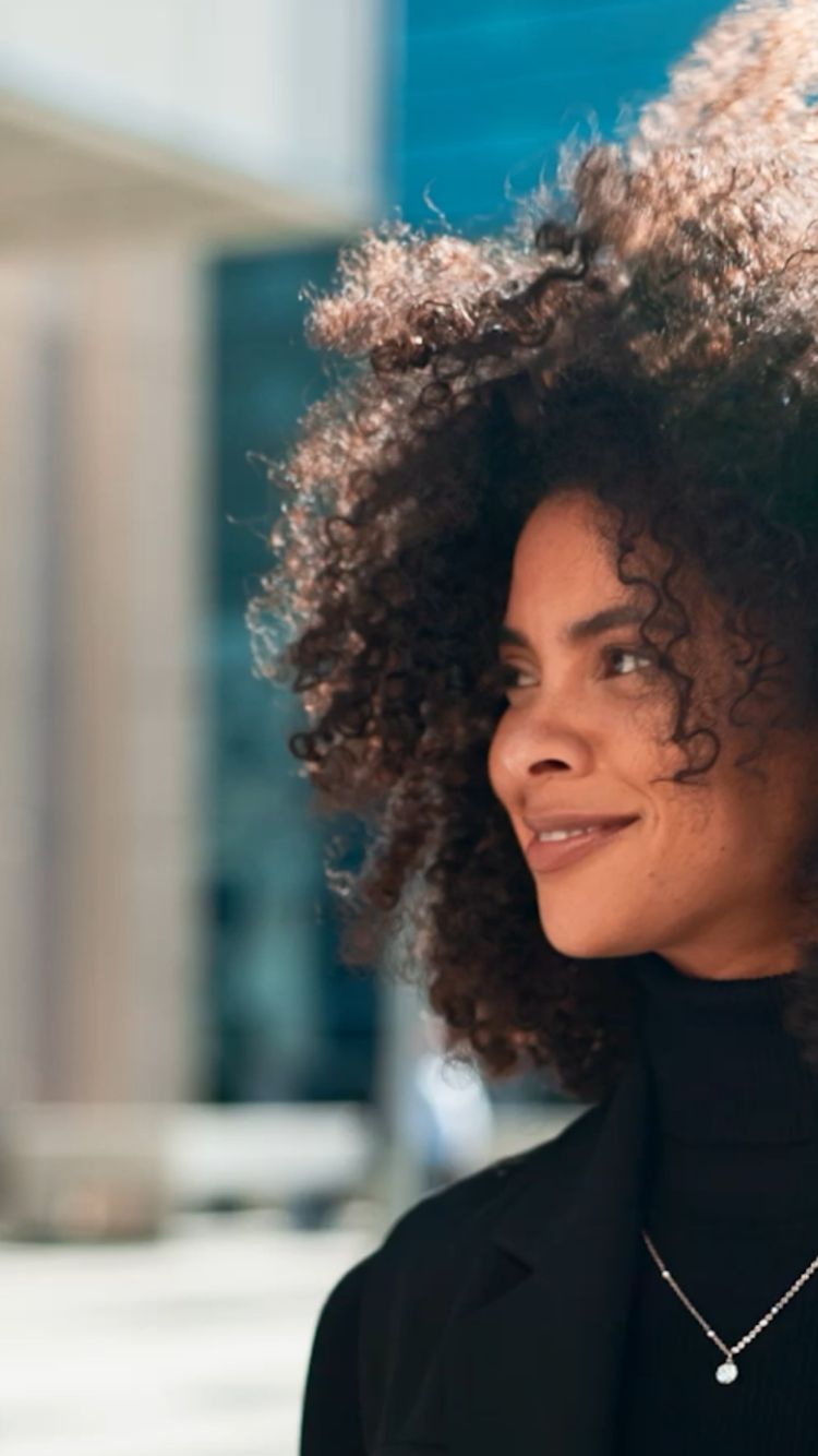 Zelfverzekerde vrouw met krullend haar, zwarte coltrui en ketting glimlacht in het zonlicht: de spirit van de FIT Group.