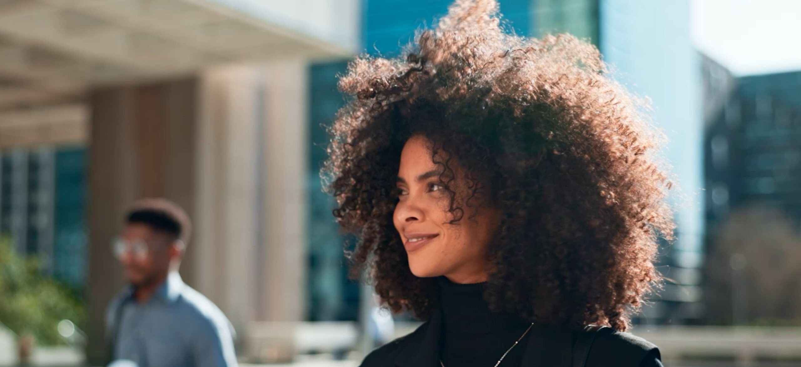 Lachende vrouw met krullend haar in de stad, glazen gebouwen en het hoofdkantoor van FIT Group op de achtergrond.