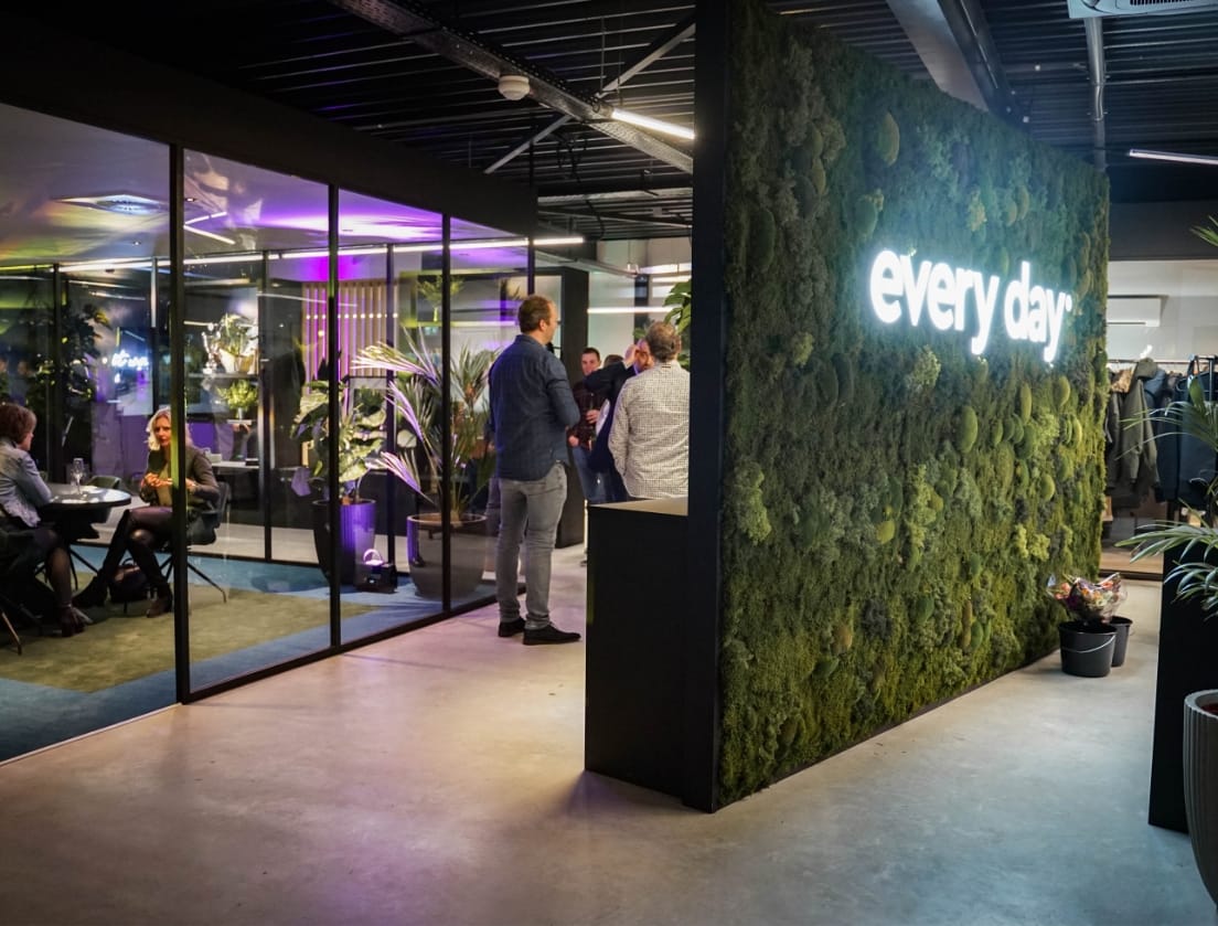 Modern Shopify B2B-kantoor: moswand met neon "elke dag"-bord, mensen die in de buurt aan het kletsen zijn.