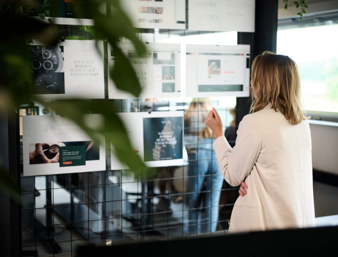 Een vrouw in een wit jasje bekijkt afgedrukte grafieken en Shopify B2B-ontwerpen op de glazen wand van haar kantoor.