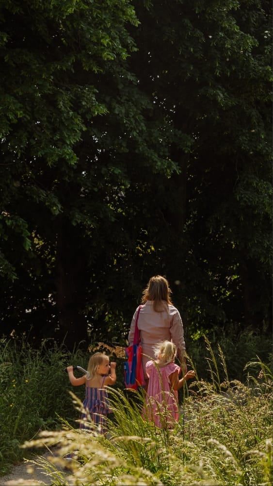 Een vrouw en twee meisjes in Memphis Depay-uitrusting lopen op een zonnige dag door hoog gras in de richting van weelderige bomen.