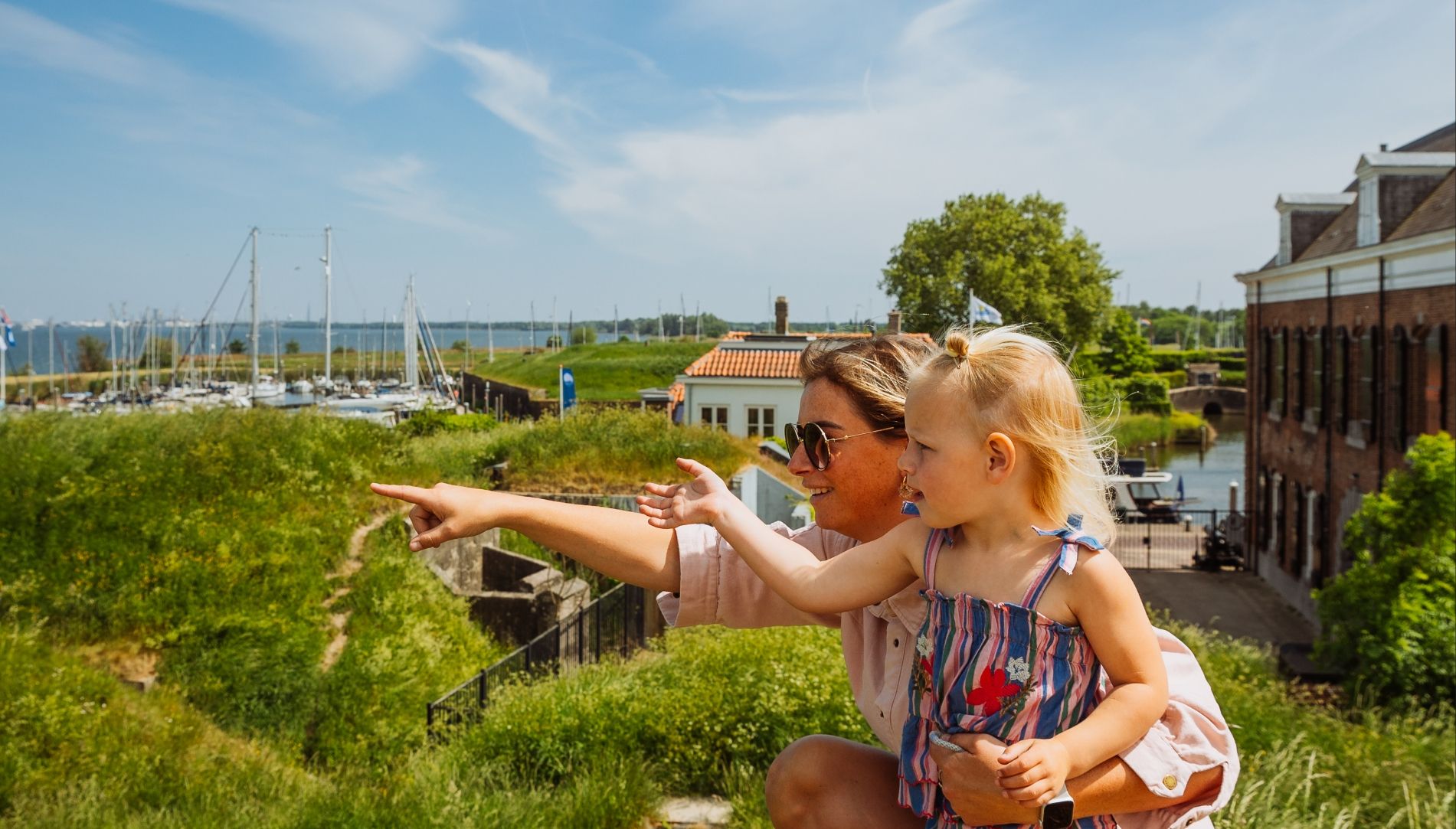 Een vrouw en meisje in Memphis Depay-uitrusting wijzen naar iets bij een zonnige jachthaven.