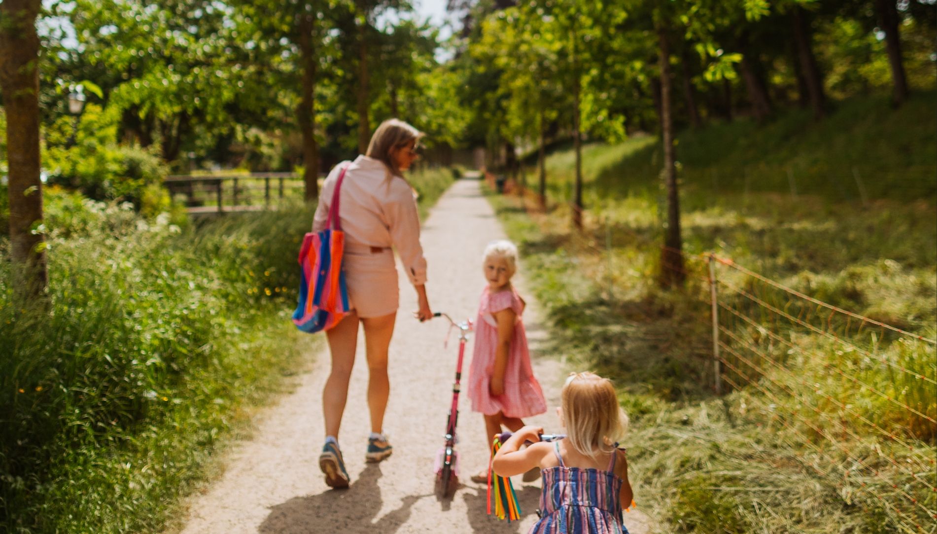 Een vrouw en twee meisjes in Memphis Depay-uitrusting lopen op een step over een zonnig, met bomen omzoomd pad.