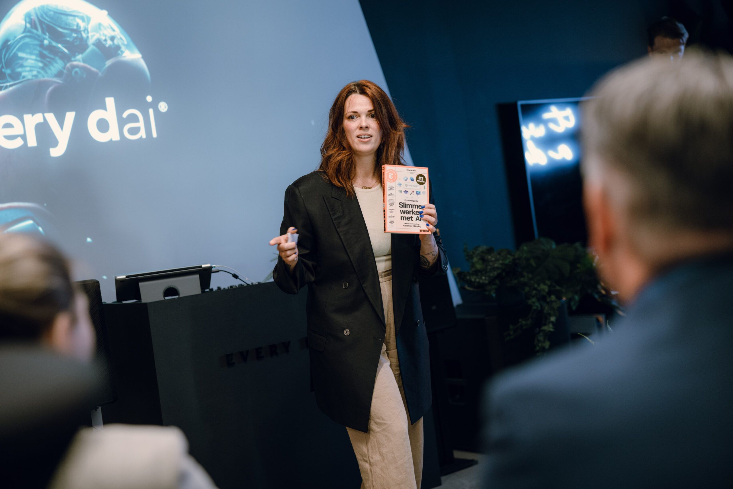 Vrouw presenteert aan een publiek, houdt een boek vast, met een presentatiescherm op de achtergrond.