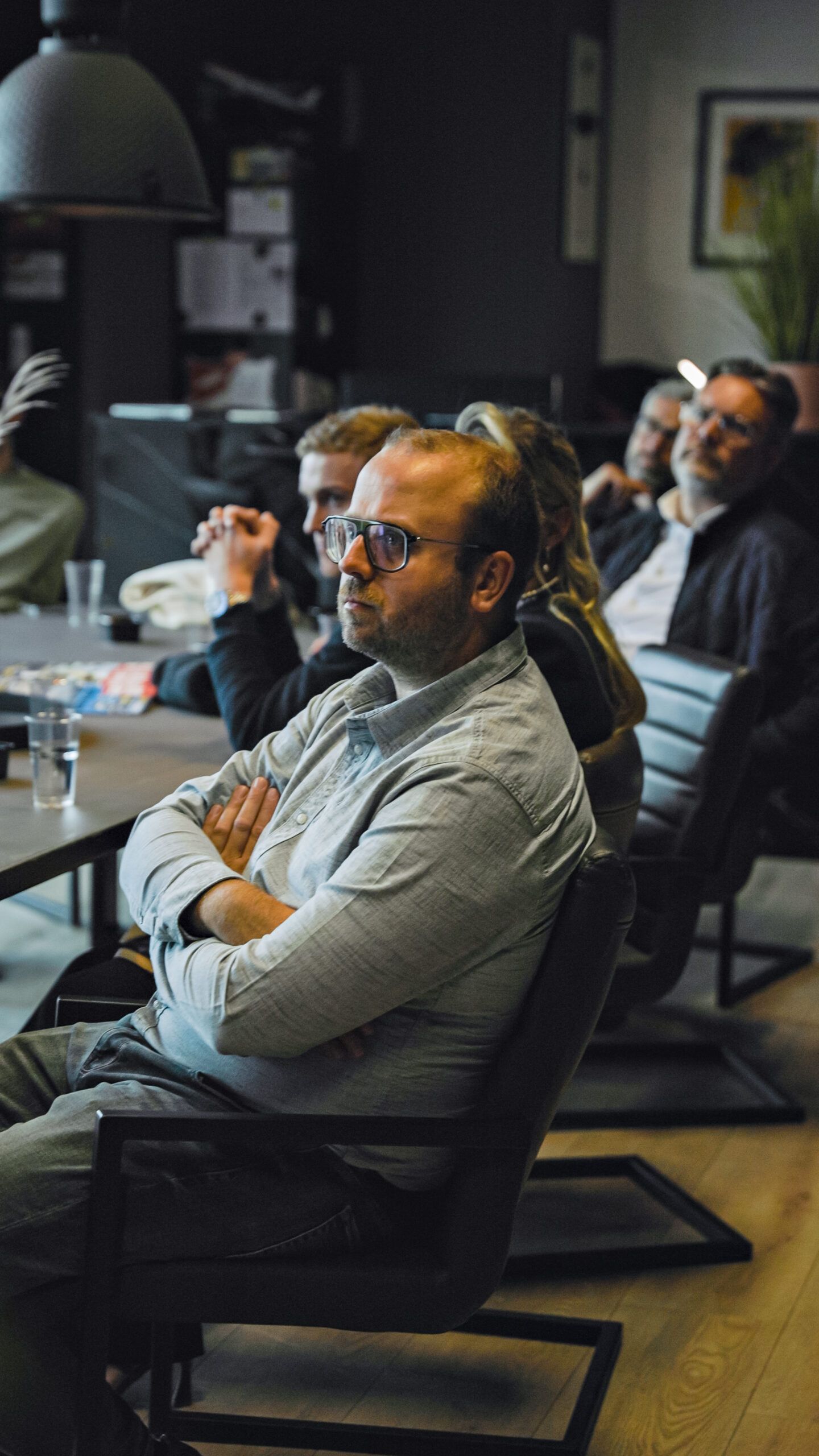 Man met bril zit met gekruiste armen te luisteren in een schemerige vergaderruimte met anderen.