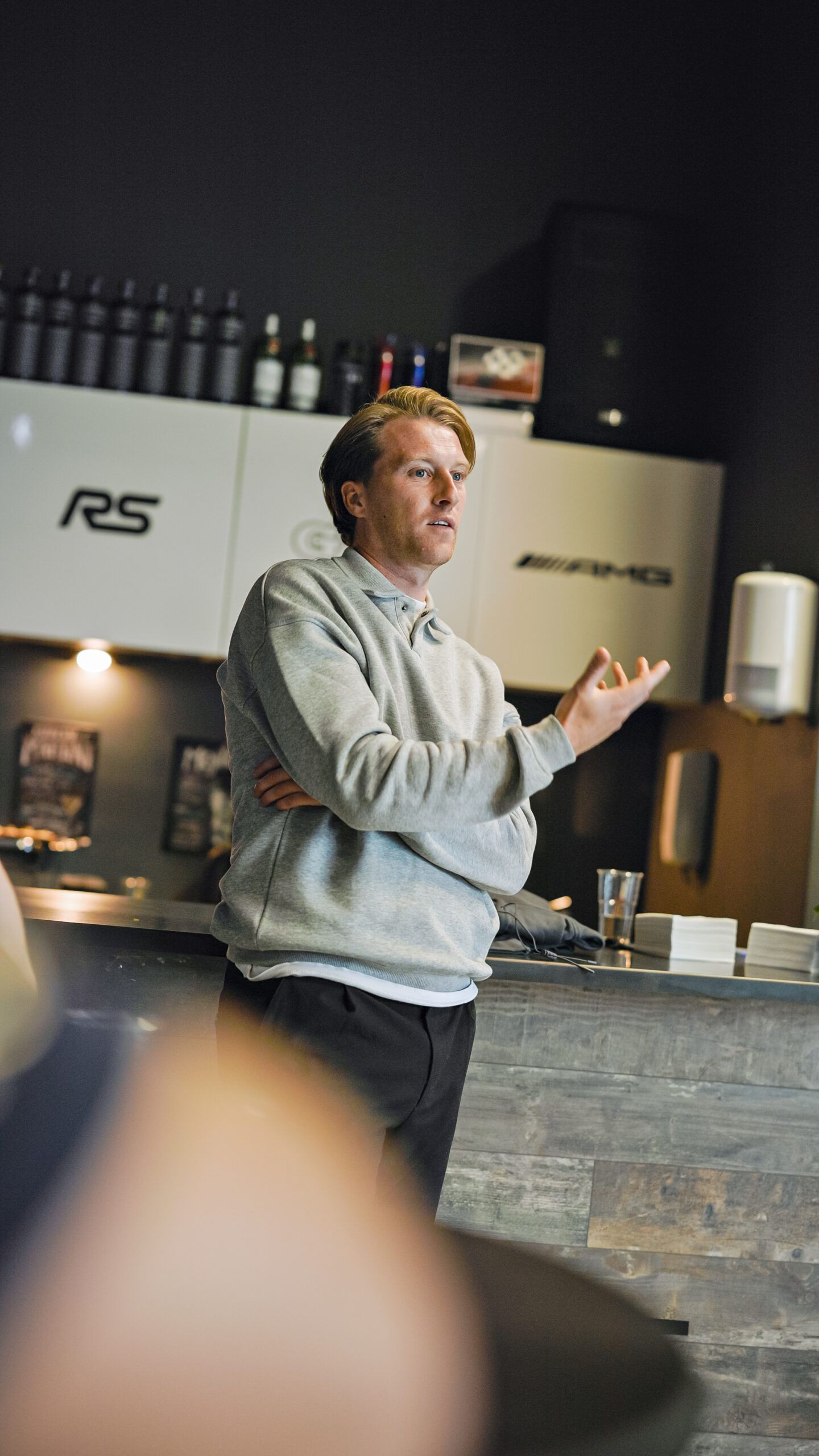 Man met grijze capuchon gebaart terwijl hij spreekt in een strakke, moderne keuken of café.