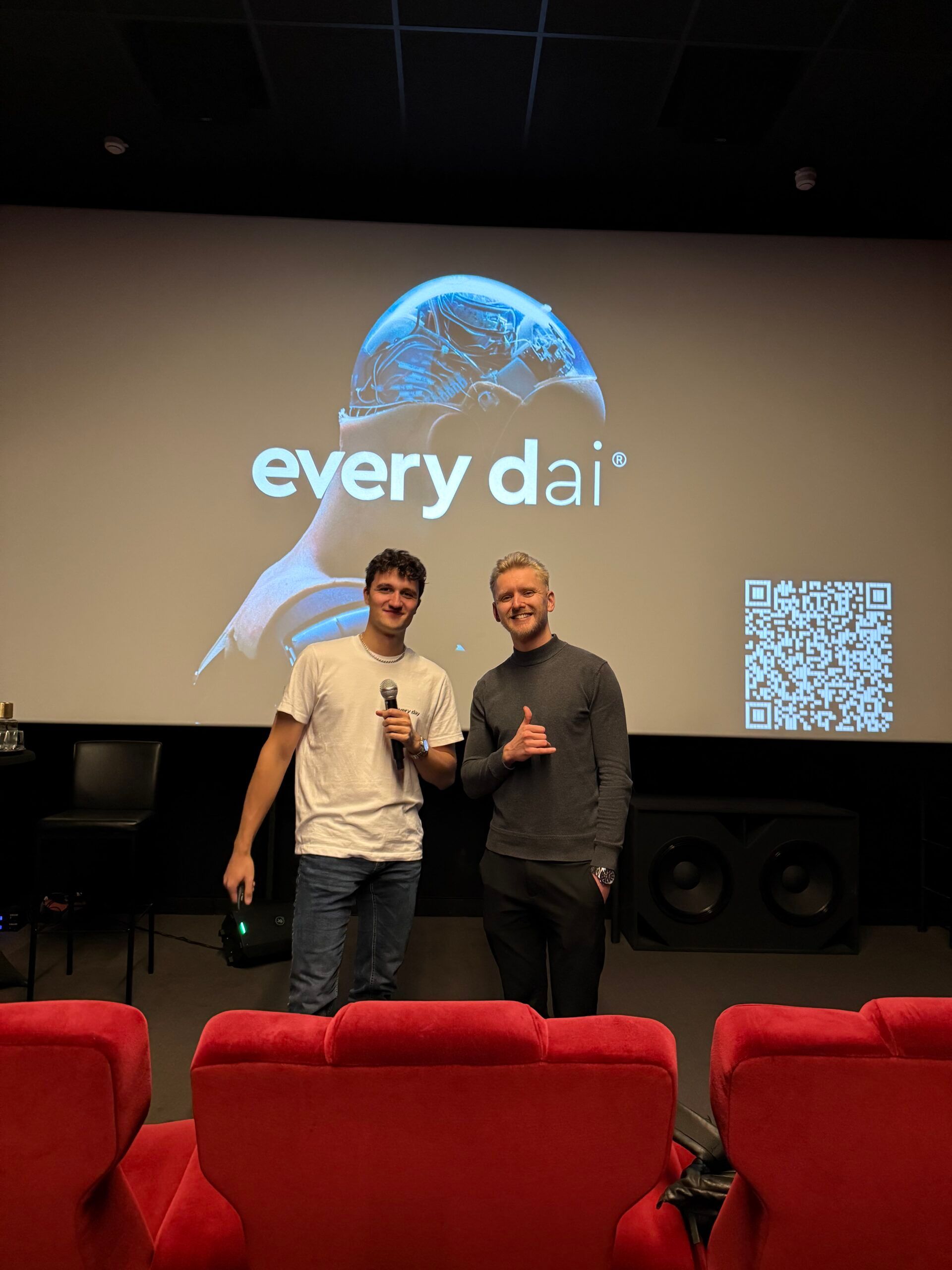 Twee mannen poseren voor een scherm met daarop elke dai en een QR-code, in een theater met rode stoelen.