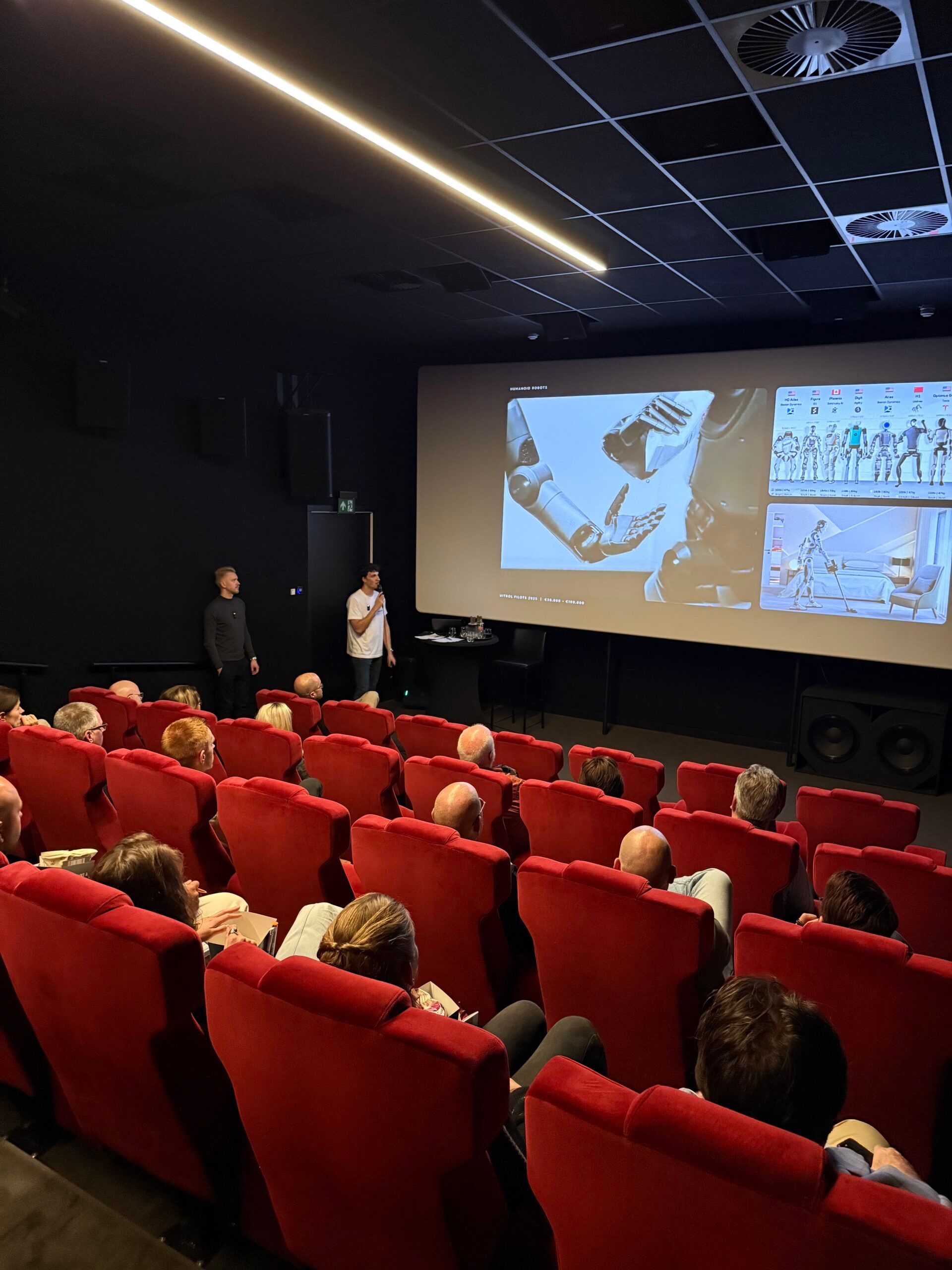 Theaterpubliek kijkt naar een techniek- en robotpresentatie op een groot scherm vooraan.