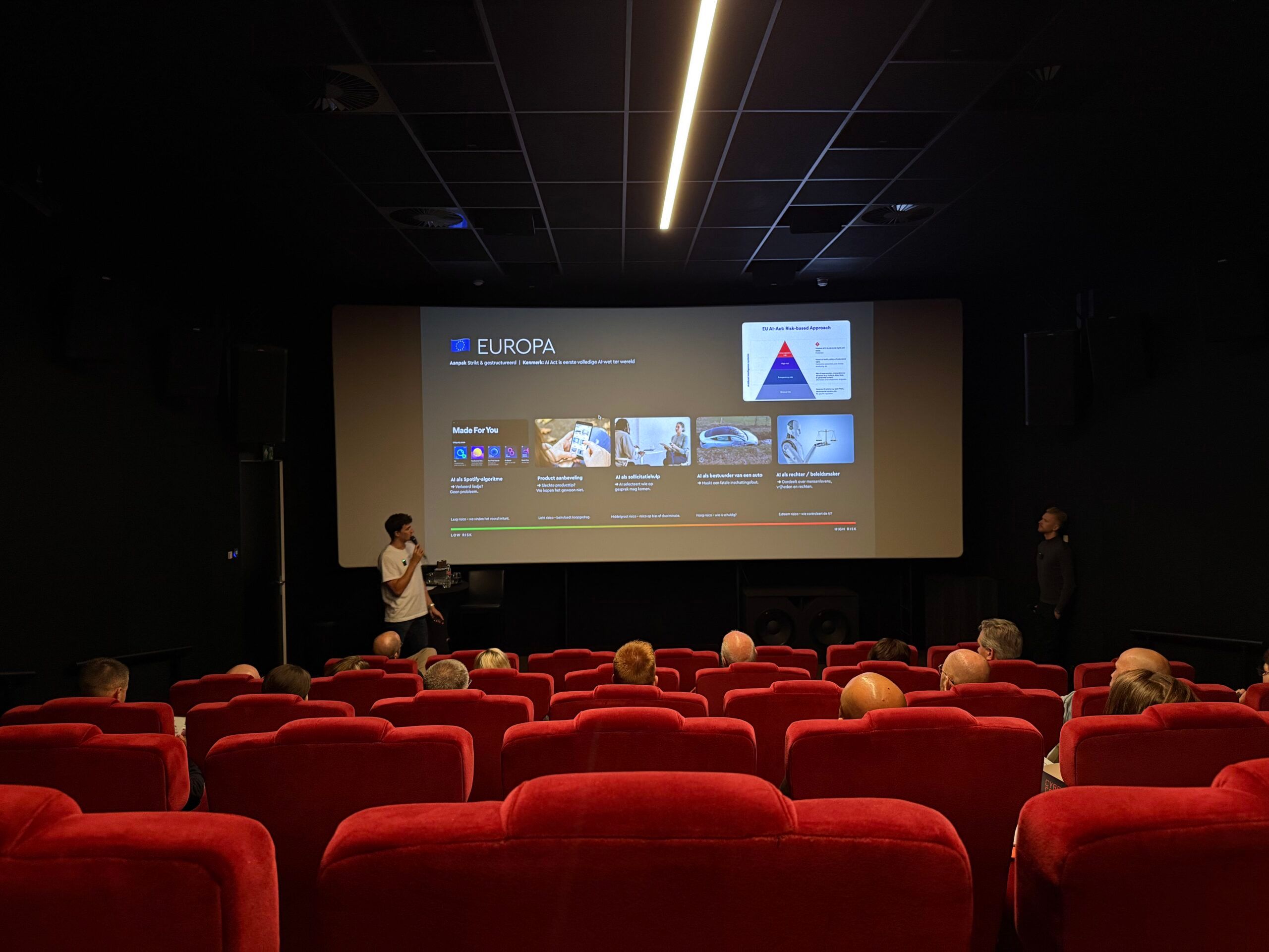 Het publiek bekijkt een presentatie op een groot scherm in een donker theater met rode stoelen.