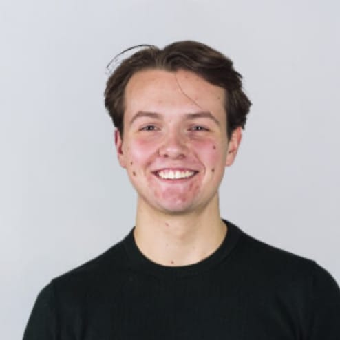 Een glimlachende jongeman met kort bruin haar in een zwart shirt staat voor een effen grijze achtergrond.