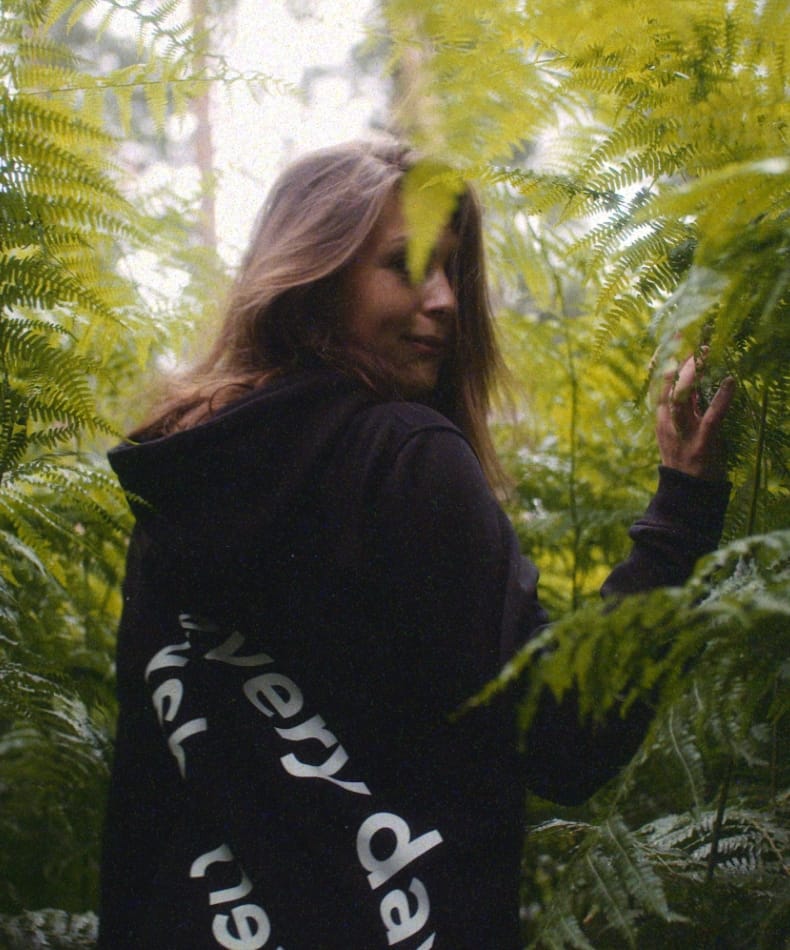 Vrouw in zwarte hoodie draait zich half om te midden van weelderige groene varens.