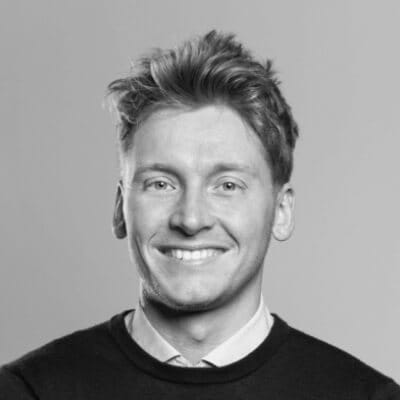 Zwart-witportret: Kortharig persoon, lachend, in een trui over een overhemd.