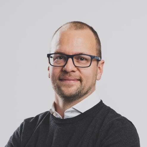 Lachende man met bril, baard, donkere trui en wit overhemd kijkt in de camera.