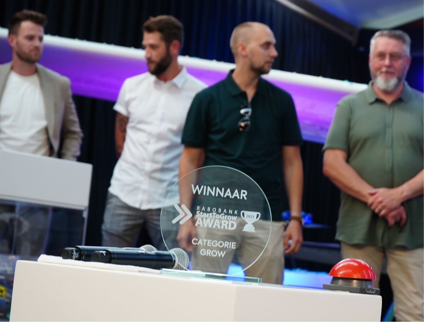 Glazen prijs op tafel naast microfoon en knop; vier mannen staan erachter.