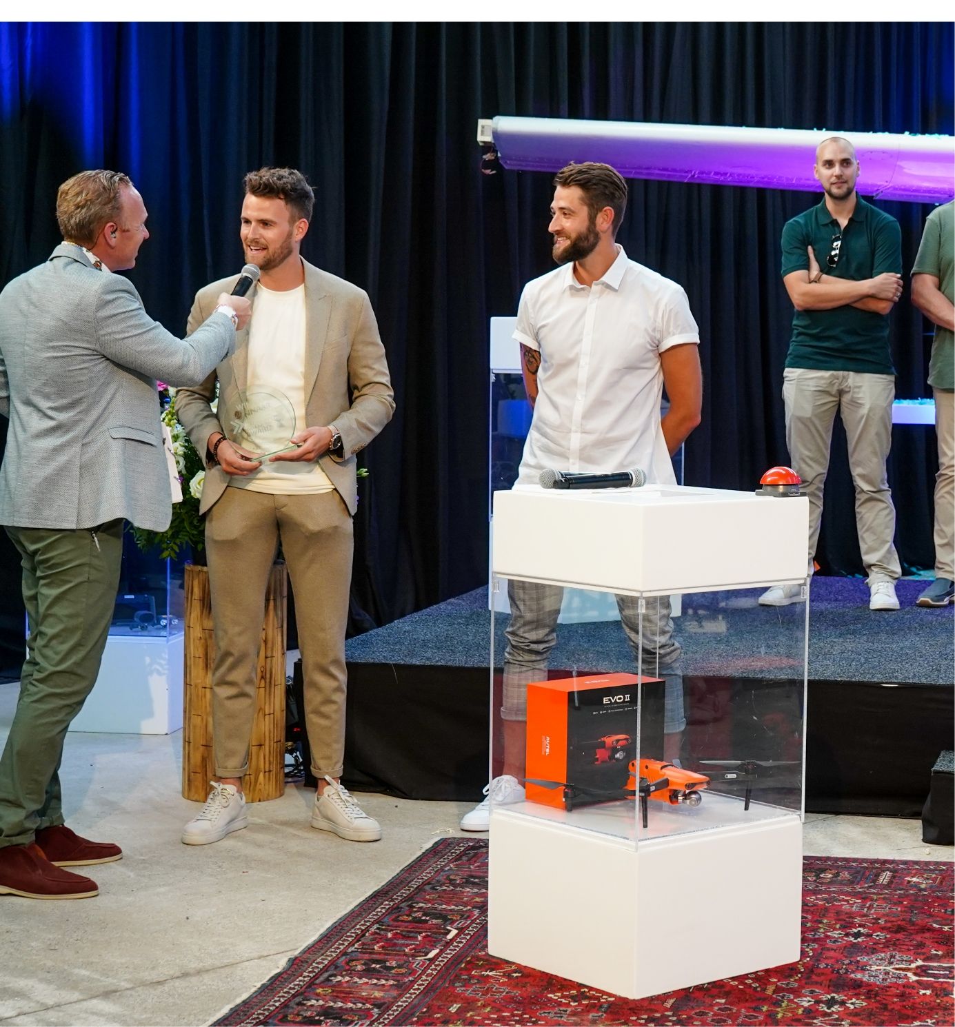 Twee mannen op het podium: een houdt een trofee vast terwijl hij geïnterviewd wordt. Een drone-show boeit het publiek.