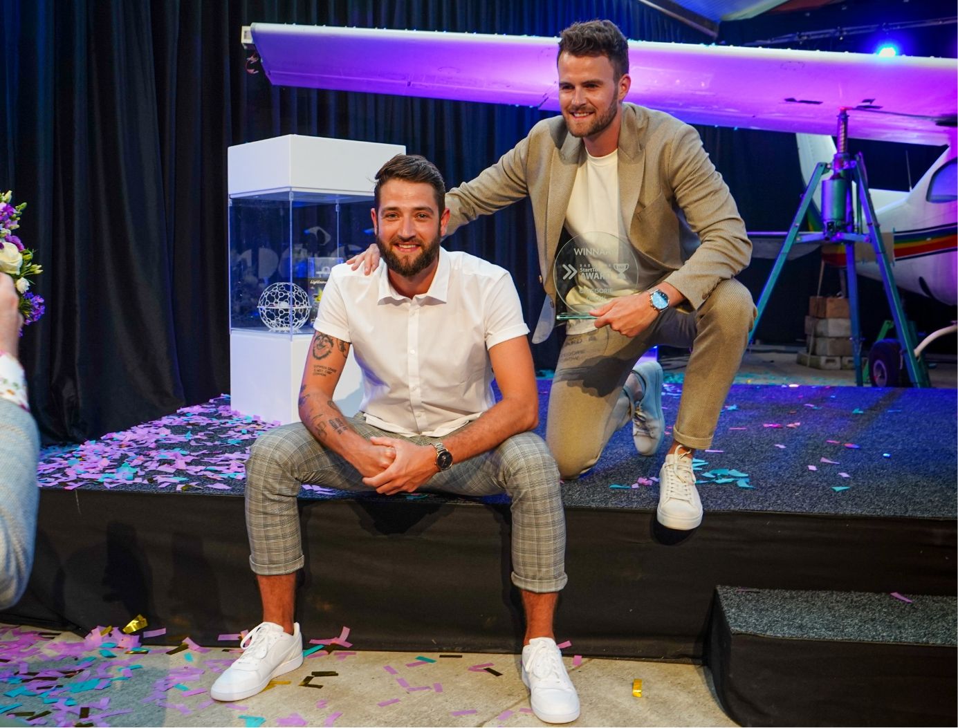 Twee mannen vieren feest op het podium met confetti; één zittend, één knielend en met een vliegtuig op de achtergrond.