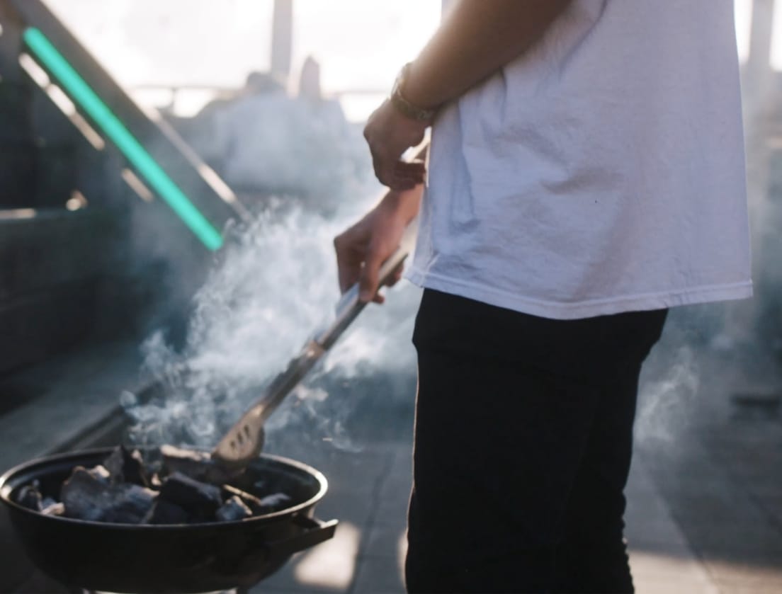 Grillen met een tang boven houtskool, gekleed in een wit shirt en een zwarte broek, terwijl de rook opstijgt.