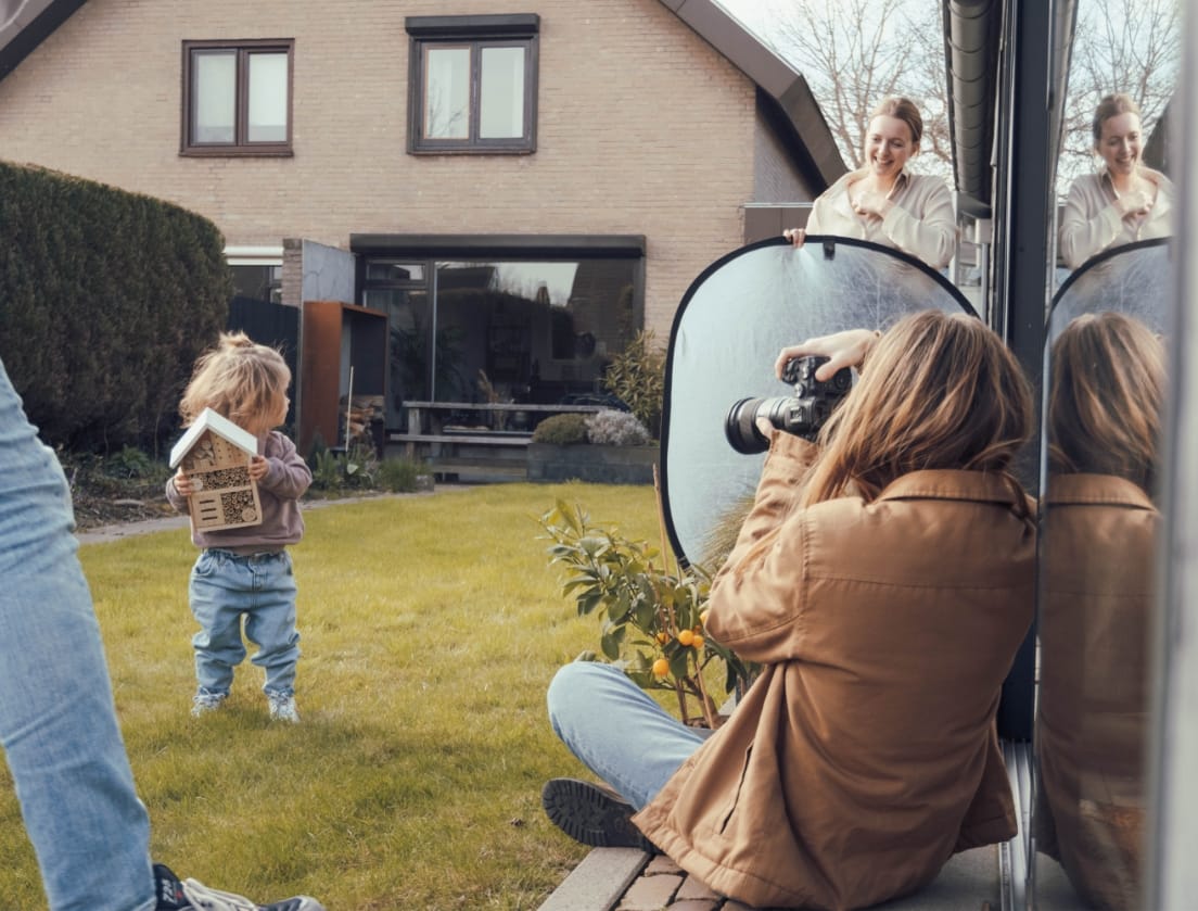 Een kind poseert met een vogelhuisje terwijl twee volwassenen een fotoshoot in de achtertuin bij een stenen huis leiden.