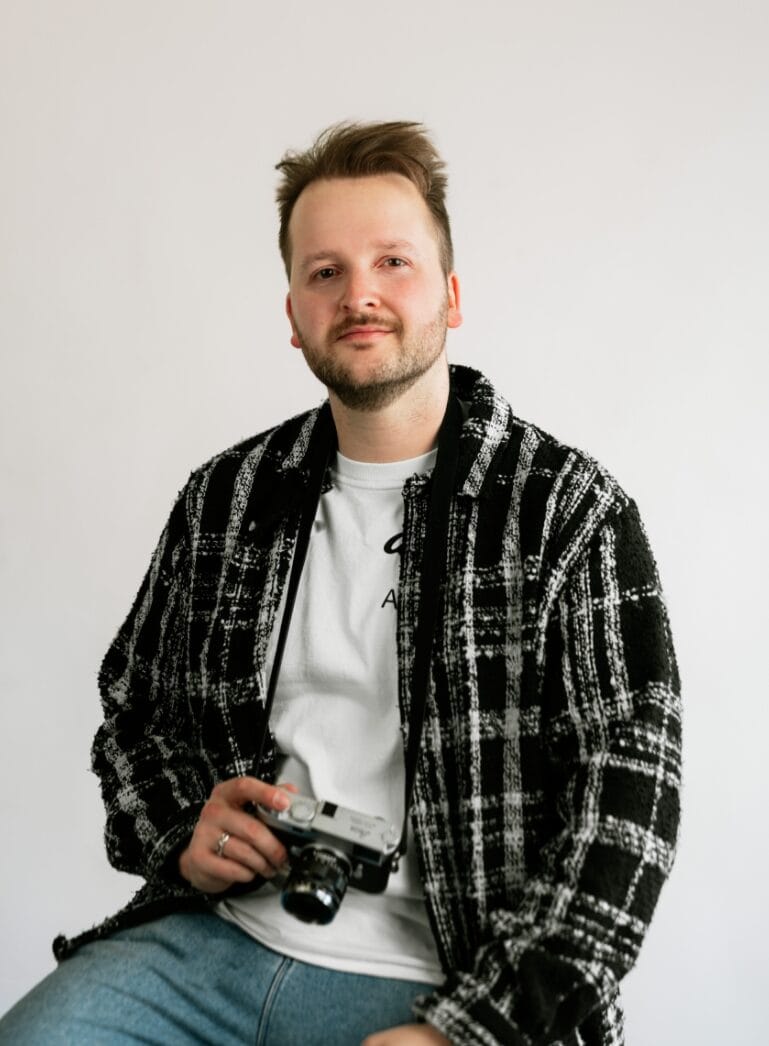 Iemand in een geruit jasje zit tegen een effen achtergrond en houdt een camera vast.