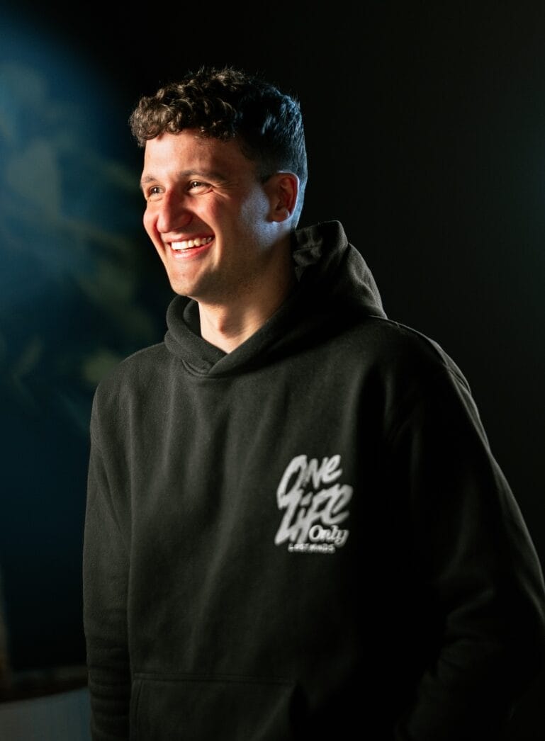 Een persoon straalt in een zwarte hoodie met de tekst 'One Life Only' erop, tegen een donkere achtergrond.