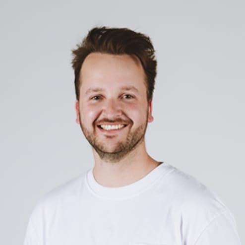 Een man met een baard in een wit overhemd, lachend tegen een eenvoudige achtergrond.