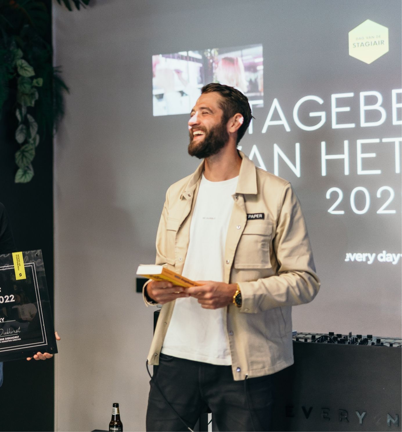 Man grijnst, houdt prijzen vast en kijkt naar een dia met daarop 'STAGIAIR 2022'.
