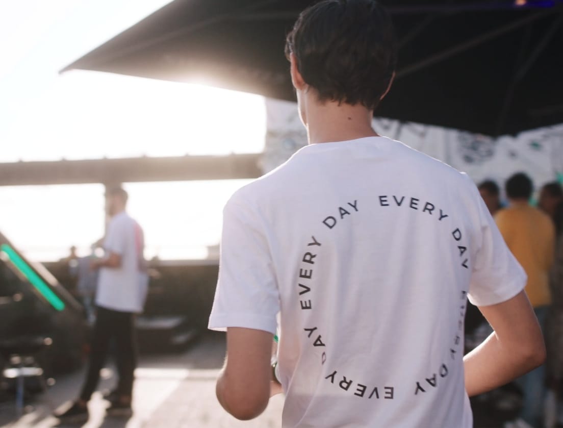 Een man in een wit EVERY DAY T-shirt loopt buiten, met zonlicht en wazige mensen op de achtergrond.