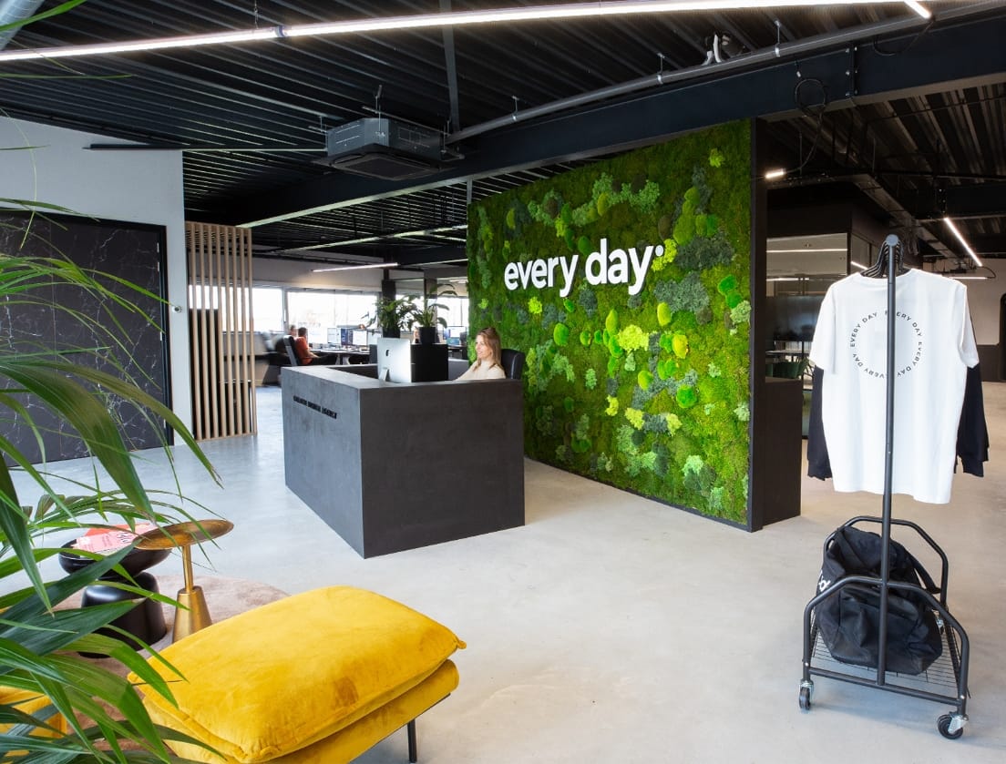 Lichte, ruime kantoorreceptie met plantenwand, logo, bureau en kledingrek.
