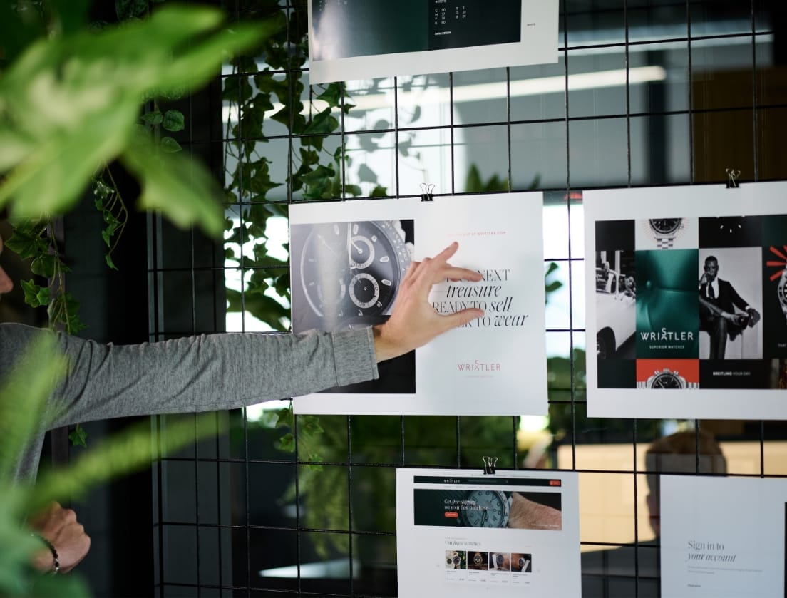 Een hand wijst naar designposters op een draadraster, omgeven door weelderige groene planten.