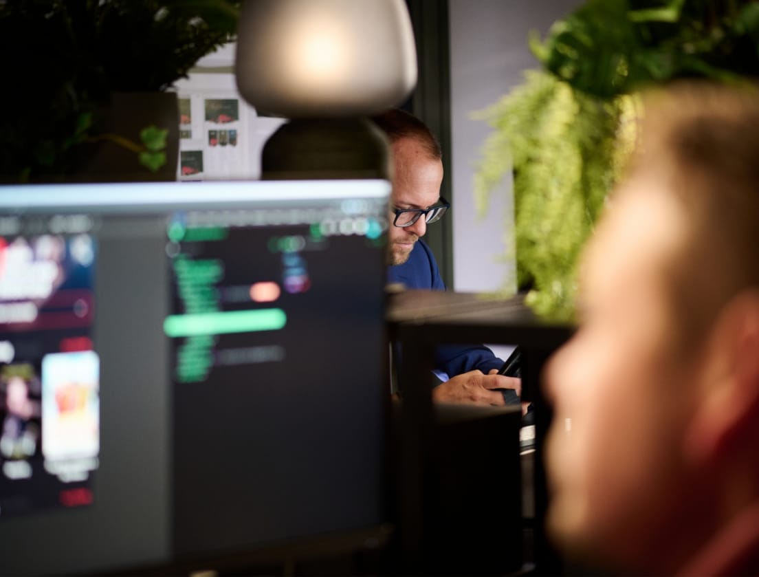 Programmeur achter computer in focus; persoon met planten vaag op de achtergrond.