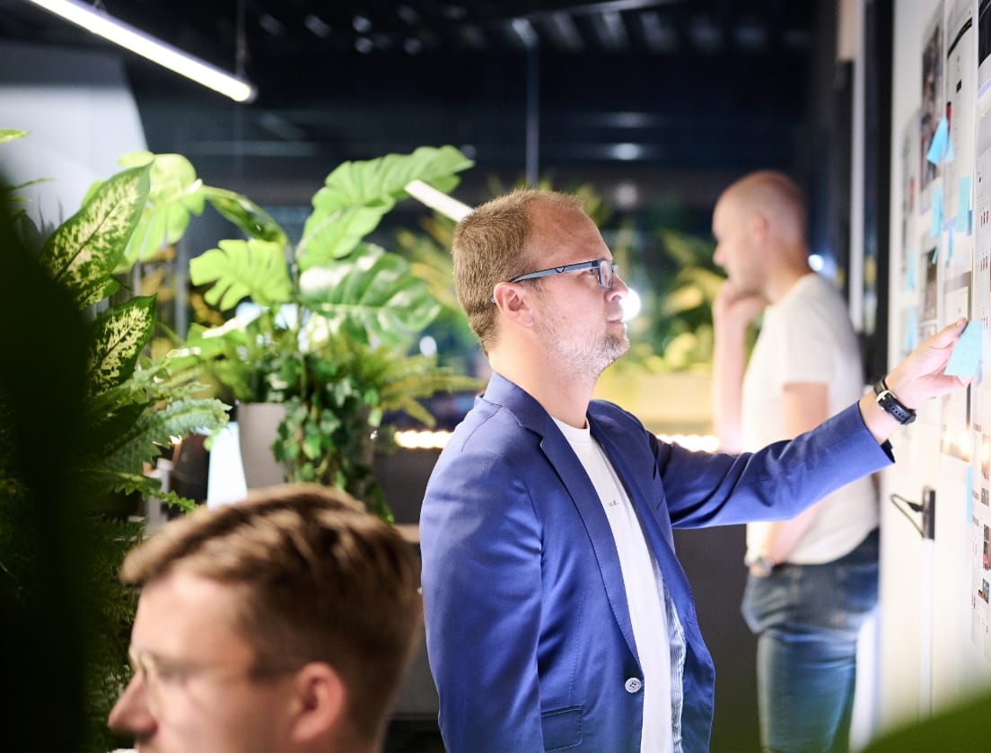 Een man in een blauw jasje schrijft aantekeningen op een bord in een kantoor vol planten.