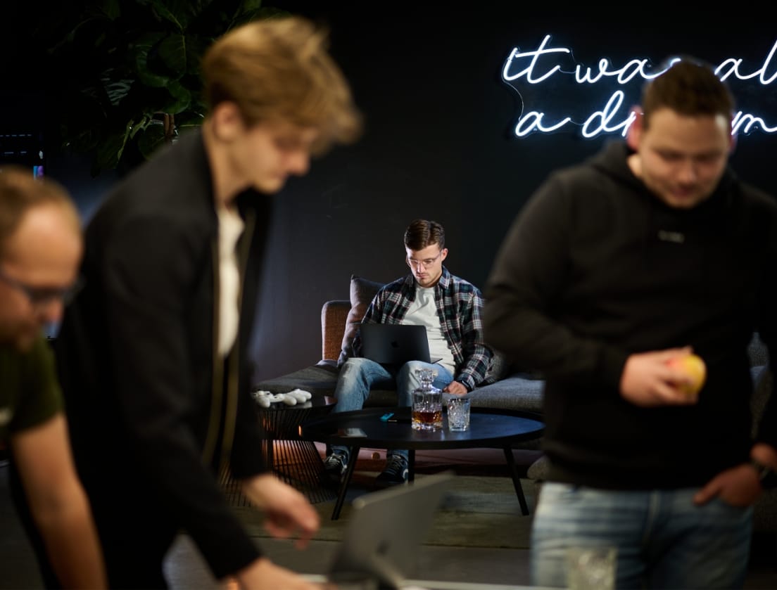 Modern kantoor: persoon achter laptop, anderen in de buurt; neonlicht op de muur.