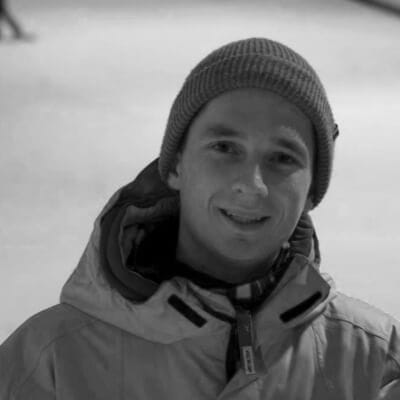 Een man in een winterjas en muts lacht naar de camera te midden van een besneeuwd landschap. Zwart-witfoto.