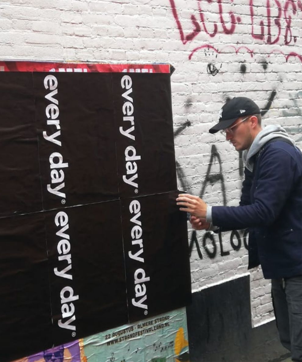 Een persoon met een pet plakt dagelijks posters op een met graffiti bedekte bakstenen muur.