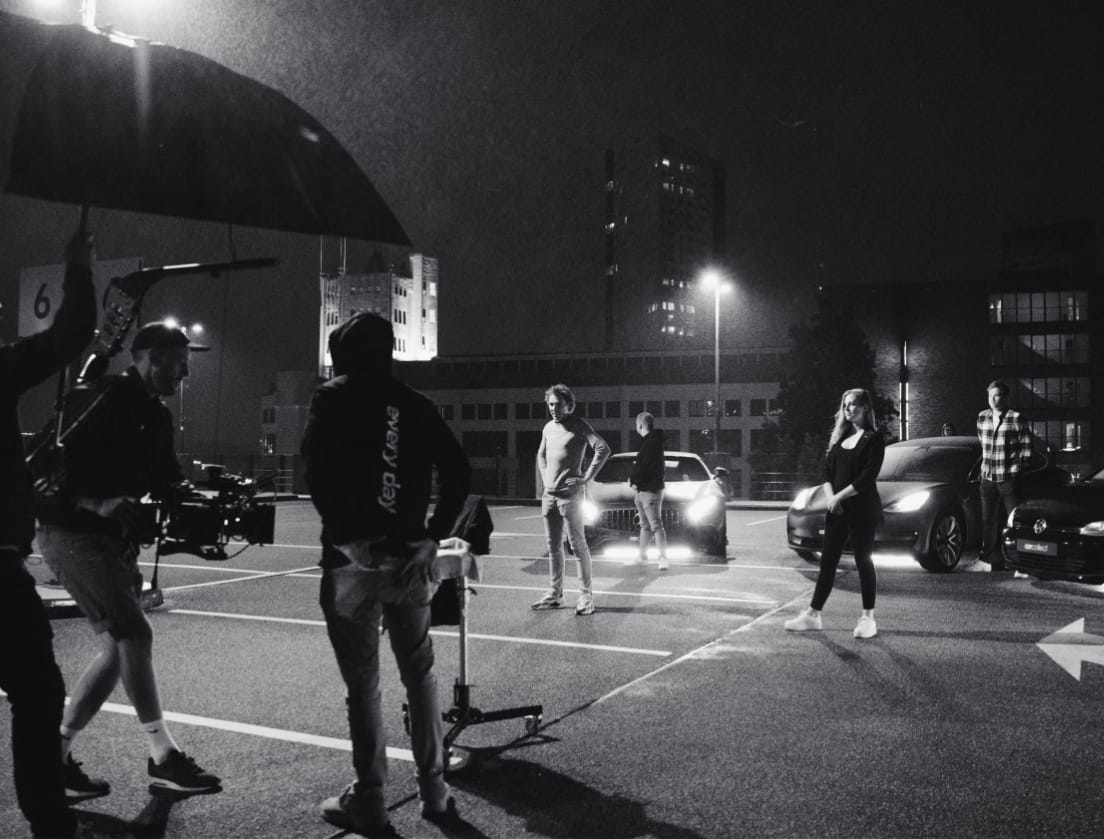 Een filmploeg filmt een nachtscène op een verlichte parkeerplaats. Acteurs en auto's zijn verlicht.
