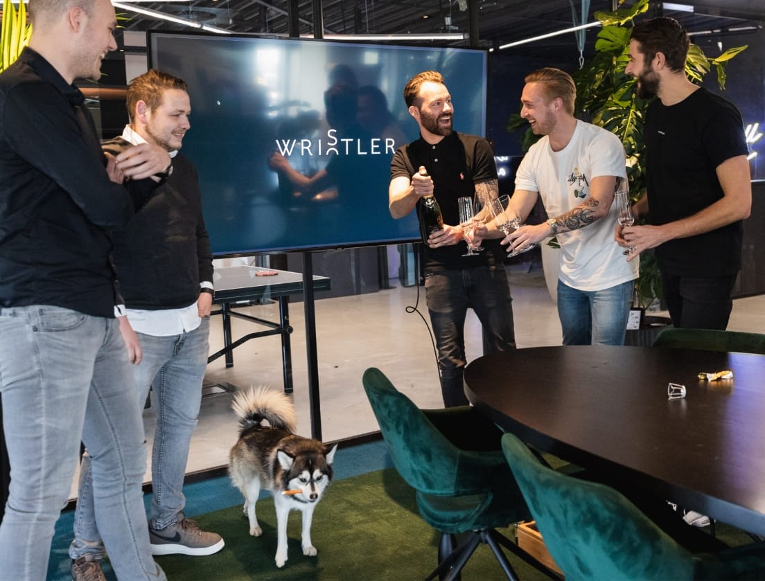 Vijf mannen toosten met champagne bij een hond en lezen op het scherm "WRISTLER" op het kantoor.