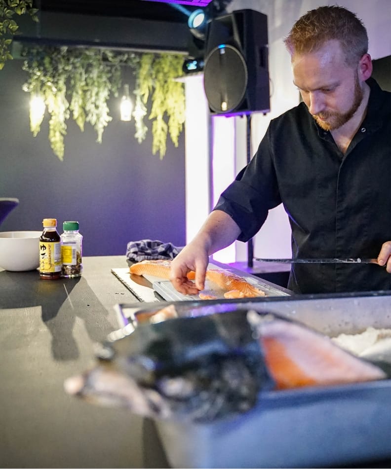 Een chef-kok in het zwart bereidt zalmfilet in een strakke keuken, omringd door hangend groen.