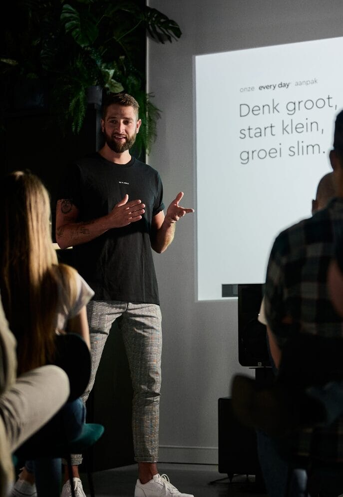 Een presentator spreekt tot een publiek; op het scherm staat: "Denk groot, begin klein, word slim.