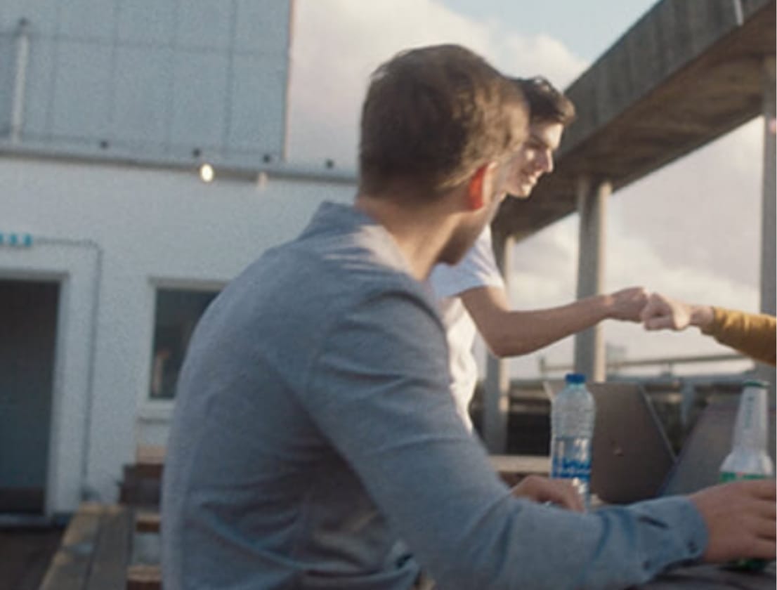 Drie op een dak: twee fist bumps, een derde in blauwe horloges. Laptops en drankjes verspreid over de tafel.