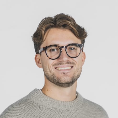 Man met bril in een beige trui, lachend tegen een witte achtergrond.