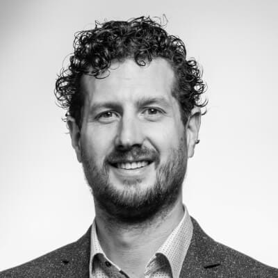 Zwart-witportret van een man met krullend haar in een blazer en overhemd, lachend tegen een effen achtergrond.