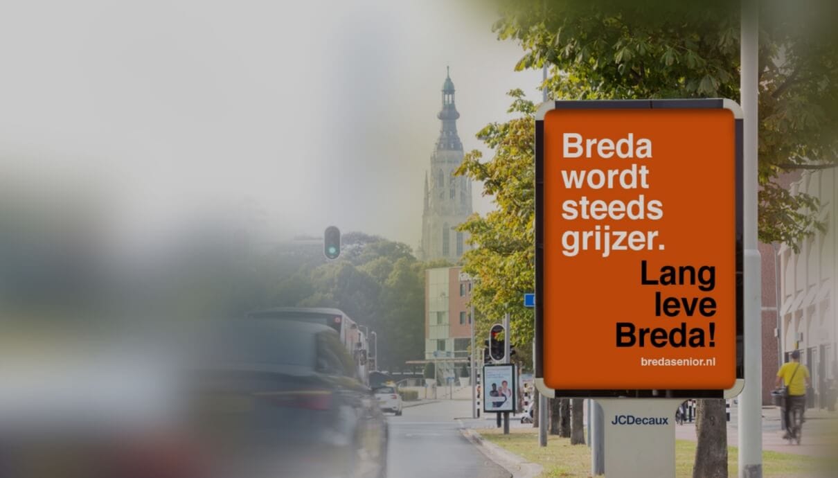 Straatbeeld: Op het reclamebord staat "Breda wordt grijzer. Leve Breda!" met een wazige kerk op de achtergrond.