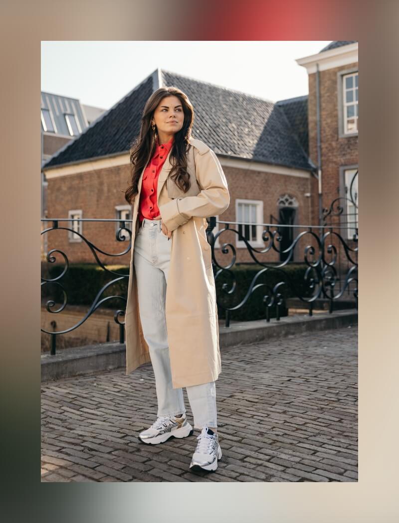 Vrouw in beige trenchcoat en jeans op een geplaveid pad langs een smeedijzeren hek.